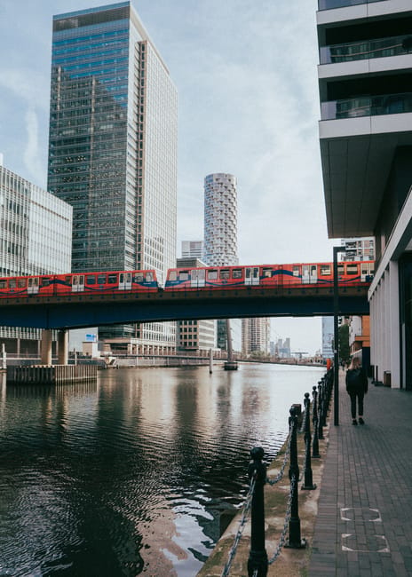 A red London Underground train is crossing a modern bridge over a river in Canary Wharf, with tall glass office buildings on either side. In the foreground, a brick-paved walkway runs alongside the water, separated by black metal bollards connected by chains. A person dressed in dark clothing is walking along the pavement, carrying a bag. To the right, part of a contemporary building with large windows and dark paneling is visible. The sky is partly cloudy, and the scene depicts an urban environment typical of a city centre area. The image relates to home relocation and furniture transport services offered by Man With a Van Docklands, highlighting the logistics of moving within this busy district.