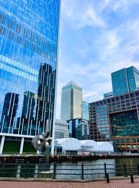 Photograph of a cityscape at Canary Wharf showing multiple modern high-rise buildings with reflective glass facades, including a large blue-tinted skyscraper on the left and a shorter, light-colored tower in the background. In the foreground, a canal with dark railing along the edge is visible, and a shiny metal sculpture is positioned near the water. The scene is well-lit with natural daylight, under a partly cloudy sky. This image illustrates the type of urban environment where tight access moves and home relocation services by Man With a Van Docklands may be required, highlighting the importance of professional furniture transport and packing during the moving process.