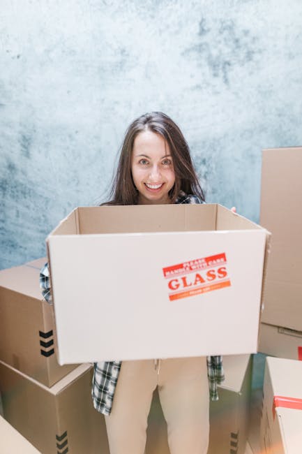 A young woman with long brown hair and a smile is standing indoors in front of a textured, light blue wall, holding an empty cardboard moving box labeled 'GLASS' with red and white packaging tape arou