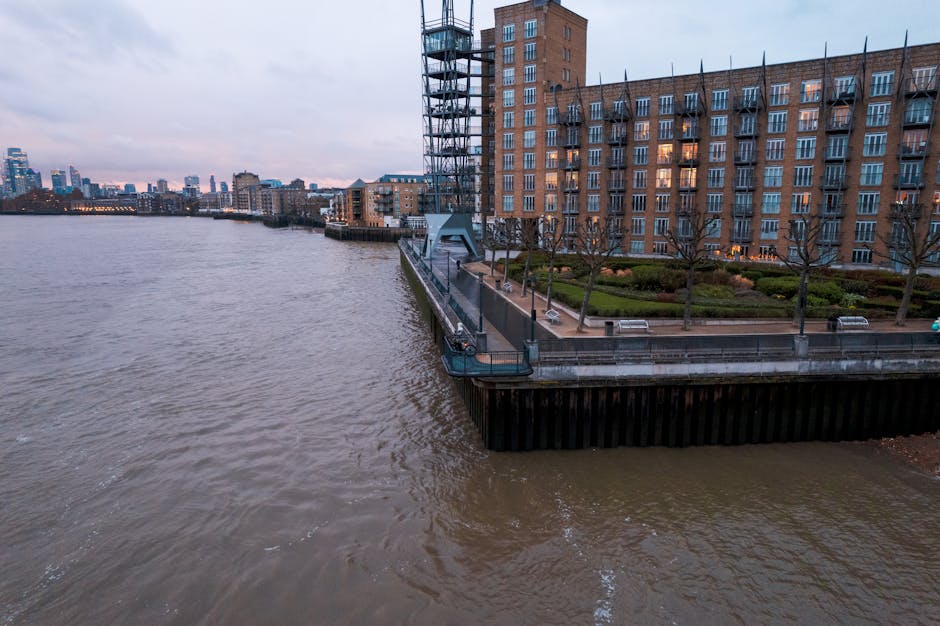 Aerial view of a riverside residential area near Mudchute station in Docklands, showing a large brick apartment building with multiple windows and a terrace with benches and small trees. In the foreground, a pontoon-style floating dock extends over the water, with a metal gangway connecting it to the pavement. The river has a brownish hue with gentle ripples, and the city skyline with various high-rise buildings is visible in the distance under a cloudy sky. The scene captures a moment of home relocation or furniture transport planning, with the company's presence subtly implied through the setting of a typical move environment involving loading or unloading near the residence in the Docklands area.