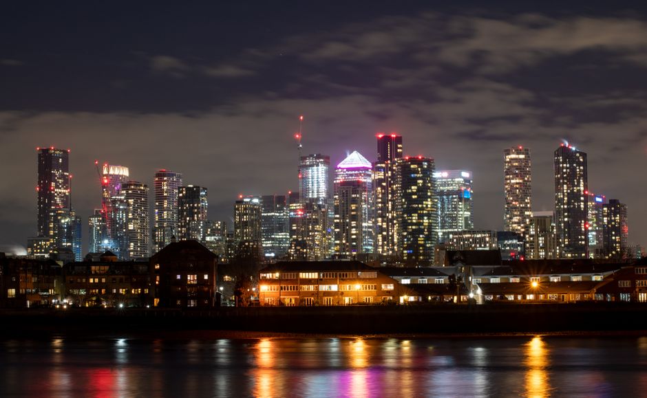 A nighttime photograph of the London Docklands skyline taken from across the River Thames. The image shows a row of illuminated skyscrapers and high-rise buildings with various brightly lit windows and modern architectural features, including antennas and spires on top. The dark sky above is partly cloudy, with some cloud cover visible. In the foreground, there are low-rise residential buildings with warm yellow lighting, and the river below reflects the city lights, creating colorful reflections on the water’s surface. This scene captures the vibrant urban environment characteristic of the Docklands area, relevant to house removals and relocation services such as those provided by Man With a Van Docklands, illustrating the area's structure and transportation context within London.