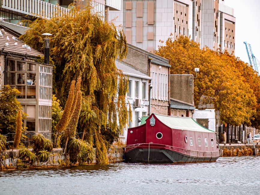 A narrow house along the Limehouse to Wapping canal features an adjacent red houseboat moored close to the water’s edge, with a leafy tree partially overhanging the boat. The scene includes residential buildings with large windows and various materials such as brick and wood, set against a backdrop of modern high-rise structures and a crane. The water appears calm, with reflections of the houseboat and trees visible on the surface. When planning a home relocation with Man With a Van Docklands, loading lorries or vans may be positioned nearby, with furniture and packed boxes being moved from the property to the vehicle. The image captures the typical urban canal environment, highlighting the challenge of narrow street access and the need for careful packing, loading, and transport logistics during furniture transport and house removals in the Docklands area.