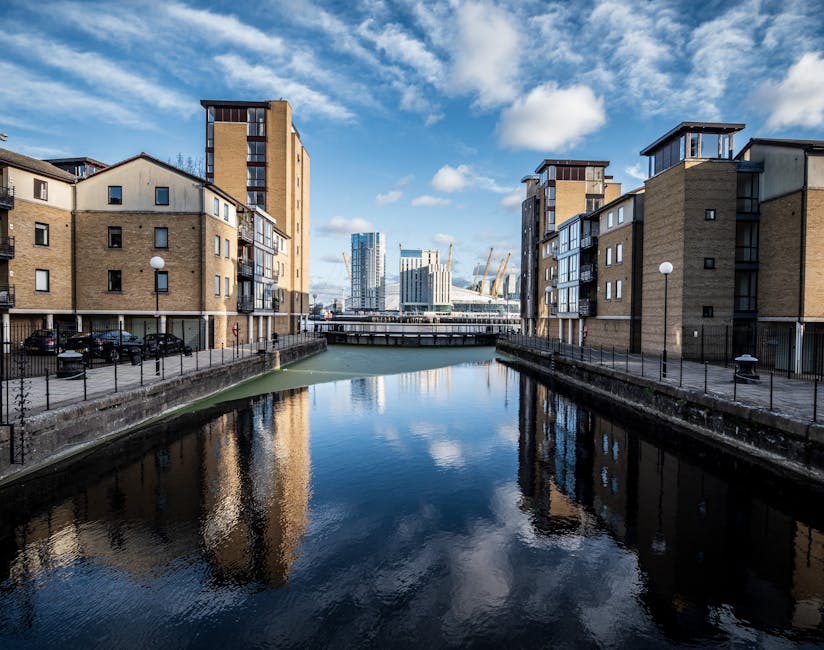 A wide view of a narrow canal flanked by modern apartment buildings constructed with beige brick and grey panel exteriors, with multiple small windows and balconies. The canal water is calm, reflecting the partly cloudy blue sky and surrounding structures. A low stone wall with black metal railings borders the canal on both sides, and there are paved walkways with evenly spaced street lamps. In the background, a bridge crosses over the canal, leading to a cluster of contemporary high-rise office buildings with glass facades and construction cranes nearby, indicating ongoing development in the Docklands area. The scene captures an urban residential and commercial environment suitable for house removals and furniture transport, with ample space for loading and unloading during home relocation or moving logistics, as coordinated by Man With a Van Docklands.