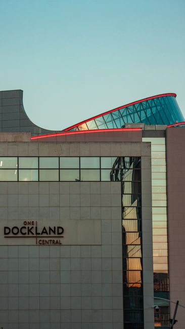 The image shows a modern multi-storey building with a curved, glass-covered roof section and a light-colored facade, featuring large windows and a sign that reads 'One Dockland Central' mounted on the
